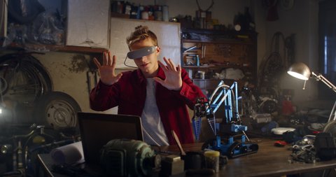 Cinematic shot of curious teenage boy with vr glasses is testing mechanical arm for innovative engineering project at school in his father workshop. 