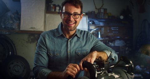 Cinematic shot of skilled man is examining and trying to fix his car engine by himself and smiling in camera in workshop.