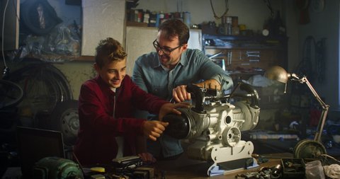 Cinematic shot of curious teenage boy and his skilled father are examining and trying to fix together car engine by themselves in workshop.