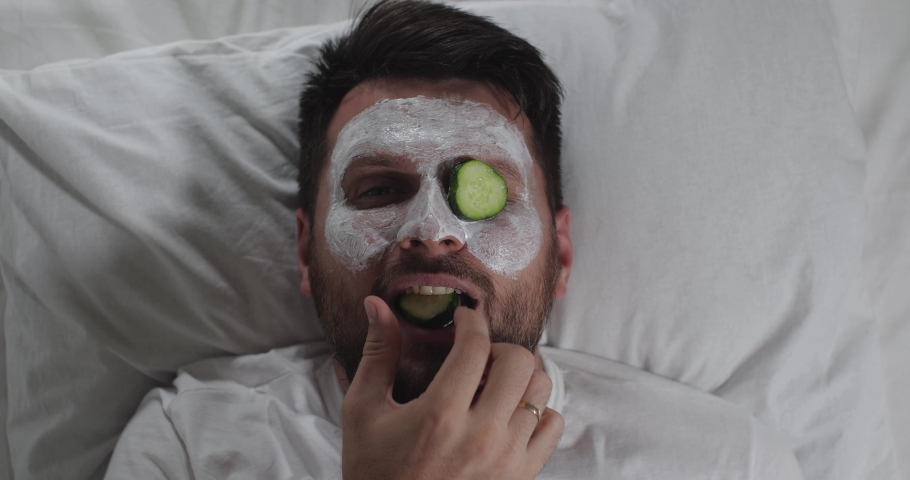 Close-up of the face of attractive bearded man lying on a couch in the beautician's office relaxing with a vitamin mask on his face. He takes cucumber slices from eyes and eats them