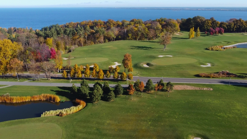 Section of park with different trees among the lawn and autumn forest located next to the big reservoir in windy sunny weather, aerial view when moving forward
