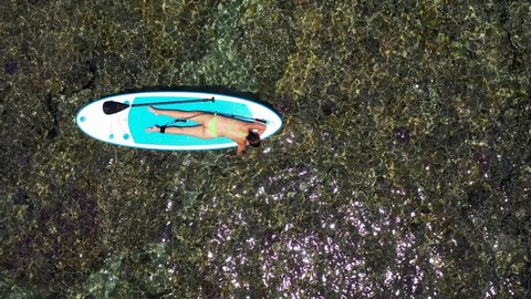 Aerial shot of a young woman lies on a paddleboard and looks at sea corals in clear water. Back view of sexy brunnette lying on a paddleboard at the blue sea