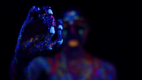 Woman painted in different luminous fluorescent paints holds pill and eats it, dark background, front view. Concept of drugs