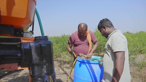 Shot of two men or male Farmers refilling chemical pesticides or fertilisers from a container into a boom sprayer vehicle or similar agriculture machine to spray in the farming field in a rural area