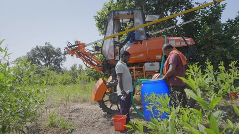 Two men or male South Asian Farmers refilling chemical fertilizers pesticides from a container into a boom sprayer vehicle or similar agriculture machine to spray in the farming field in a rural area