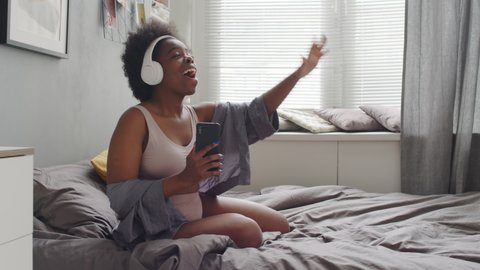 Medium slowmo shot of young happy plus size African American woman in casual underwear dancing and singing alone in bedroom while listening music in headphones