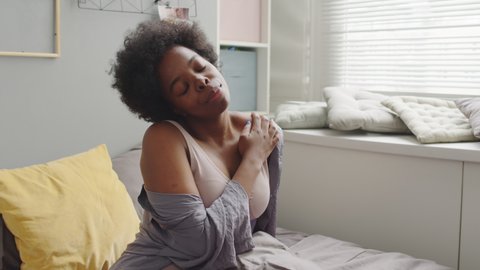 Medium slowmo portrait of young relaxed African-American woman in cozy homewear awakening in bed on weekend morning, stretching and looking at window