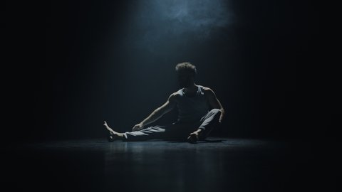 Handsome Isolated Ethnic Male Dancer Sitting on the Floor of an Empty Dark Theatre Room, Stretching His Muscles Before Starting The Workout To Prepare for the Dance Recital Performing at the Theatre.