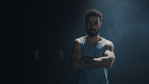 Isolated Handsome Ethnic Male Dancer With a Beard Coming Out of Smoke in a Dark and Hazy Theatre Room, Smiling and Crossing His Hands on His Chest, Looking Directly at the Camera, Spotlight on Him.