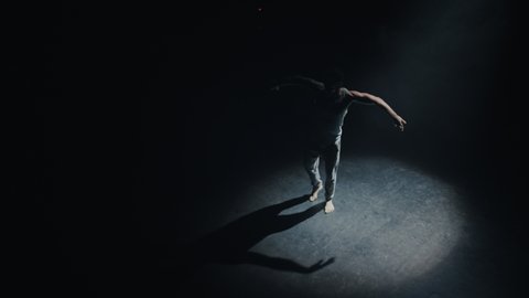 Handsome Isolated Ethnic Male Dancer Dancing Alone in the Middle of an Empty Dark Theatre Room With Spotlight Shining On Him, Wearing a Blue Tank Top and Sweatpants. Male Ballerina Practising.