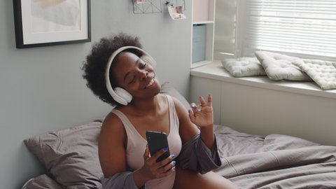 Medium slowmo shot of cheerful young African-American woman in casual pink underwear dancing to music in headphones sitting on bed at cozy bedroom