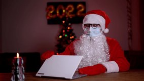 Young bearded Santa Claus wearing face mask on xmas eve sitting at cozy home table late in night talking via laptop video call. Christmas 2022 during Covid 19 coronavirus social distance concept.