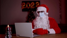 Young bearded Santa Claus wearing face mask on xmas eve sitting at cozy home table late in night talking via laptop video call. Christmas 2022 during Covid 19 coronavirus social distance concept. - Powered by Shutterstock - Get 15% off with code: PIKWIZARD15