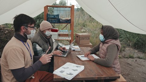 Medium shot of couple of social workers in face masks with laptop and digital tablet sitting under tent in poor refugee camp talking to immigrants, sitting down at their table one by one