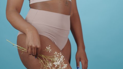 Midsection shot of unrecognizable plus size African-American woman in pink underwear sliding beautiful babys breath flower over her curvy body standing on blue background