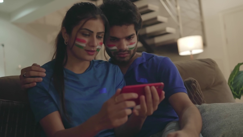 A young couple watching and enjoying a cricket match on a mobile or smart phone.  Indian Husband and wife with cheeks painted with tricolor flag intensely watching a hockey game together in a home  Royalty-Free Stock Footage #1082414335
