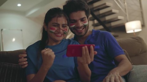A young couple watching and enjoying a cricket match on a mobile or smart phone.  Indian Husband and wife with cheeks painted with tricolor flag intensely watching a hockey game together in a home 