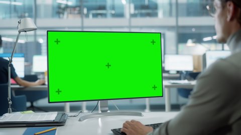 Close Up Over the Shoulder Shot of a Businessman Working on Desktop Computer with Chroma Key Green Screen Mock Up Display. Digital Projects Manager Typing Data, Using Keyboard and Mouse.