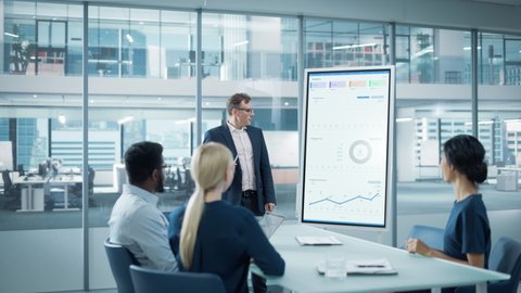 Company Operations Manager Holds Meeting Presentation for a Team of Economists. Adult Male Uses Digital Whiteboard with Growth Analysis, Charts, Statistics and Data. People Work in Business Office.