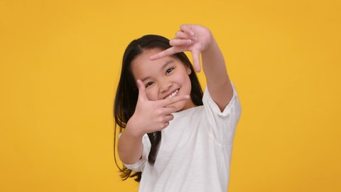 Smile to camera. Cute little asian girl making frame with fingers and smiling, pretending to make photography, orange studio background, slow motion