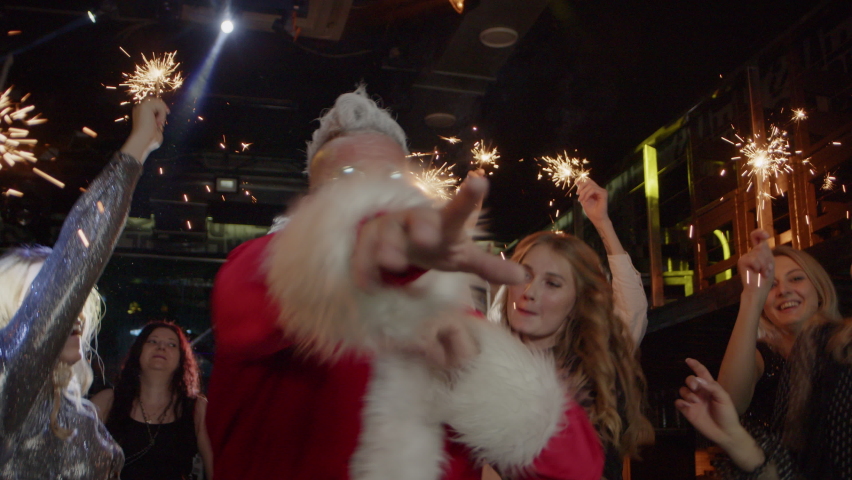 Happy smiling crowd of young people together with Santa Claus haves fun, actively dances holding magical sparklers at costume party on New Year Eve indoors. Winter holiday celebration to modern music Royalty-Free Stock Footage #1082419402