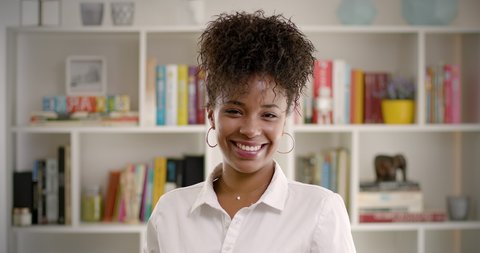 Happy Millennial Authentic Real Afro Female Close up Front Portrait. Slow Motion of Beautiful Confident Young African American Woman Looking at Camera Smiling in Front of Bookshelf. 