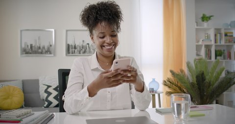 Cheerful Young African American Woman Using Smartphone at Home Office. Black Smiling Woman Using App on Cellphone. Beautiful Girl Smiling and Thinking While Chatting on Mobile Phone. Lateral Dolly.