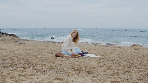 Blonde woman is sitting on blanket, enjoying her picnic on ocean beach, reading interesting book, taking pleasure of her lifestyle, young and successful, Zoom In, Slow motion.
