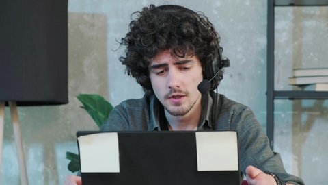 Handsome young curly haired man using laptop computer with headset to talking online with their friends or working at support line, man is trying to explain something.