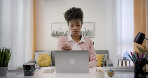 Dolly Out of Confident and Cheerful African American Woman Opening and Typing On  Laptop, Young Female Professional Writing an Important Email From her Home in a Modern Living Room. Remote Working.