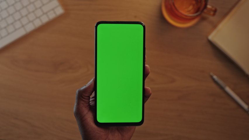 Top view of black male hands holding vertical mobile phone with green mock up screen. African american man using modern smartphone at workplace. Royalty-Free Stock Footage #1082429851