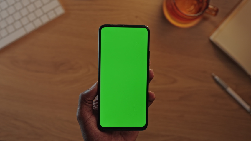 Top view of black male hands holding vertical mobile phone with green mock up screen. African american man using modern smartphone at workplace. Royalty-Free Stock Footage #1082429851