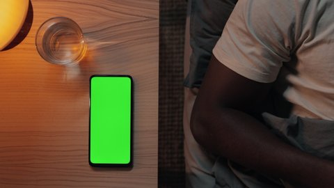 Top view of african american man resting in cozy bed and tapping on green mockup screen of modern smartphone that placed on table. Close up, chroma key.