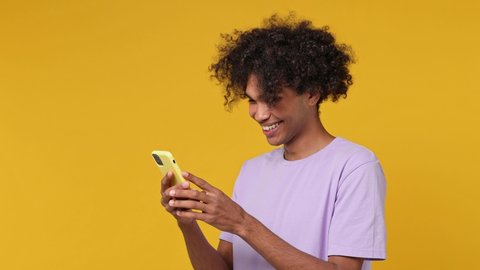 Satisfied fun young curly african american man 20s years old wears white t-shirt hold use mobile cell phone typing browsing swipe do online shopping isolated on plain yellow background studio portrait