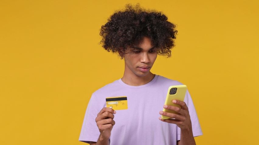 Young curly african american man 20s wears white t-shirt using mobile cell phone hold in hand credit bank card doing online shopping booking tour isolated on plain yellow background studio portrait Royalty-Free Stock Footage #1082429944