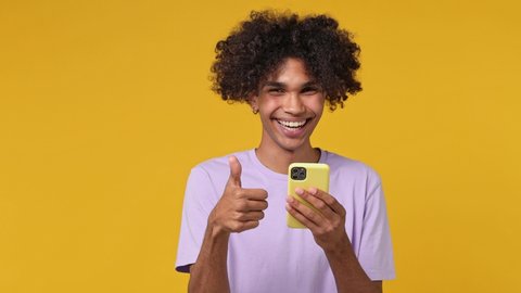 Fun young curly african man 20s wears white t-shirt hold use point index finger on mobile cell phone typing say wow yes just found out big win news isolated on plain yellow background studio portrait