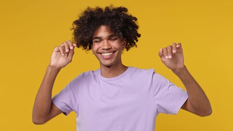 Smiling young curly african american man 20s wears white t-shirt dance clenching fists waving rising gesticulating hands have fun enjoy celebrate isolated on plain yellow background studio portrait