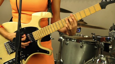 Beautiful girl plays the electric guitar. Musician in a bright orange dress. Designer stringed musical instrument of light color. Recording in a professional music studio. Perfect acoustic sounds