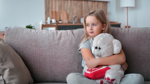 Upset lonely baby blonde girl hugging teddy bear toy sit on couch home living room interior. Unhappy female kid suffering solitude with soft plaything. Charity, adoption, waiting parents, orphanage