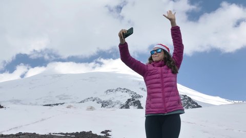 Happy woman blogger in Santa hat and sunglasses dances and shows to subscribers snowy mountain via phone standing on hill top