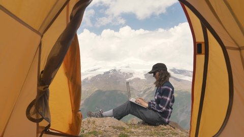 Young woman tourist in shirt opens laptop and types sitting on flat rock against large mountains on sunny day view from inside tent
