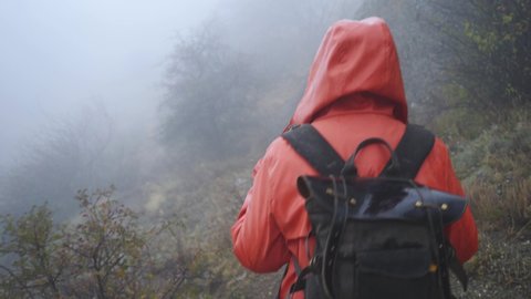 Woman in red raincoat with backpack goes on pathway in misty mountain in cold autumn day backside view, tracking shot