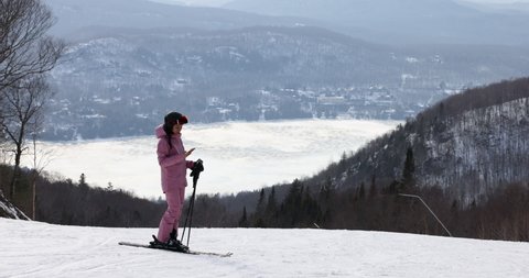 Phone on ski vacation - Woman skier using phone app on ski trail slope in amazing winter nature landscape. Girl looking at mobile smartphone wearing awesome ski clothing, helmet and goggles
