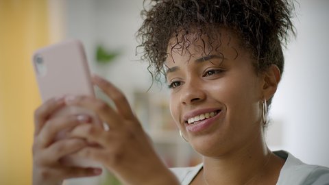Lateral Dolly of Cheerful Young African American Woman Using Smartphone at Home or Office. Black Smiling Woman texting on Cellphone. Beautiful Girl Chatting on Mobile Phone.