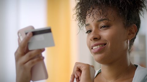 Close up Slow Motion of Smiling Young African American Woman Holding Credit Card and Smartphone at Home. Happy Female Shopper Using Mobile Phone to Make a Purchase, Online Shopping. 