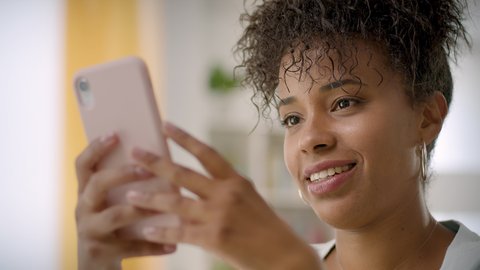 Lateral Dolly of Cheerful Young African American Woman Using Smartphone at Home or Office. Black Smiling Woman typing on Cellphone. Beautiful Girl Chatting and Laughing.