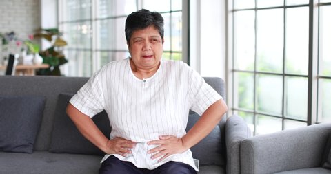 Senior 60s Asian woman suddenly feels stomach ache and use hand hold body with suffering during stay lonely in home living room. Concept of body health problem of old people.