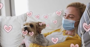 Animation of hearts moving over caucasian woman wearing face mask holding her pet dog. spending time alone at home during covid 19 pandemic digitally generated video.