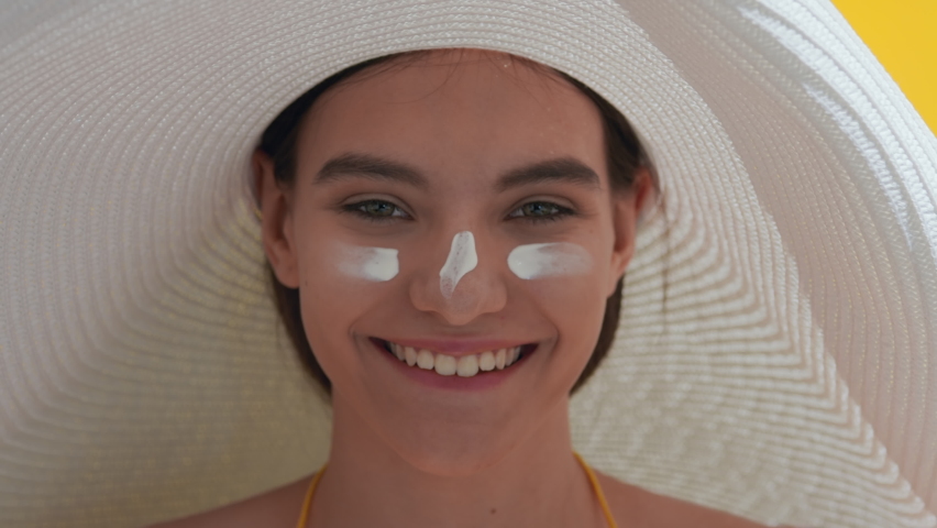 Close-up beauty portrait of happy young attractive dark-haired Caucasian woman in a big white hat with spf cream on her cheeks and nose laughing against yellow background | UV protection concept | Shutterstock HD Video #1082456542
