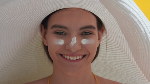 Close-up beauty portrait of happy young attractive dark-haired Caucasian woman in a big white hat with spf cream on her cheeks and nose laughing against yellow background | UV protection concept